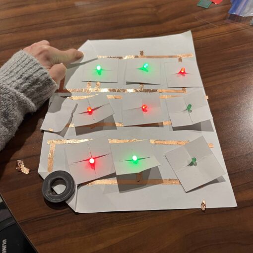 A paper tic tac toe board that features red and green LED lights connected to copper tape. The circuits are closed, which cause the lights to light up